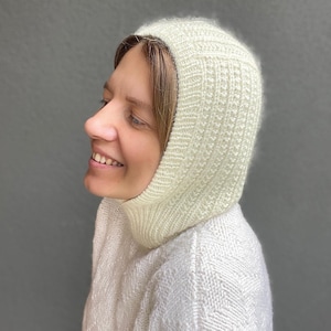 May include: A white knitted balaclava with a fuzzy texture. The balaclava is worn by a person with brown hair and is pulled up to cover their ears and neck.