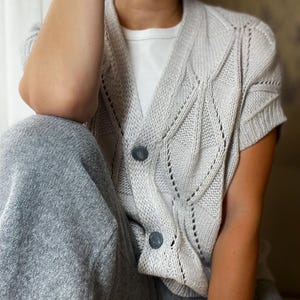 Puede incluir: Un cárdigan de punto gris claro con mangas cortas y dos botones grises oscuros. El cárdigan tiene un patrón de diamantes y se usa sobre una camiseta blanca. La persona también lleva un pantalón de chándal gris.