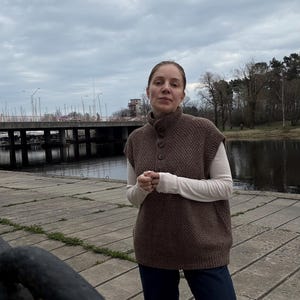 May include: A woman wearing a brown knitted sleeveless sweater with a white long-sleeved shirt underneath, blue jeans, and a neutral expression. She is standing on a concrete surface near a body of water.
