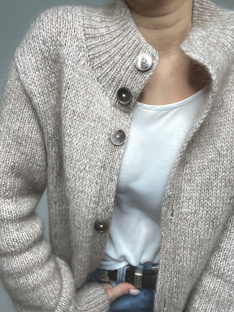 May include: A beige knit cardigan with a round collar and large, round buttons. The cardigan is made of a soft, textured yarn. The buttons are a darker shade of beige. The cardigan is worn over a white top.