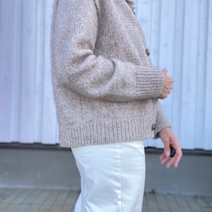 May include: A beige cardigan sweater with a ribbed hem and cuffs. The sweater has a button-up front and is made of a soft, textured knit material. The model is wearing white pants. The overall style is casual and comfortable.