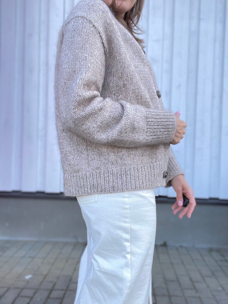 May include: A beige cardigan sweater with a ribbed hem and cuffs. The sweater has a button-up front and is made of a soft, textured knit material. The model is wearing white pants. The overall style is casual and comfortable.