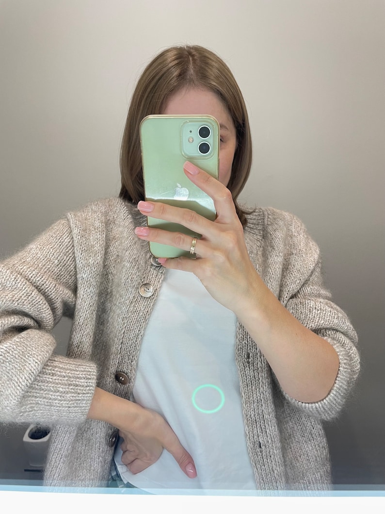 May include: A light beige cardigan with large buttons is worn over a white t-shirt. The person is holding a light green phone in front of their face. A gold ring is visible on the person's finger.