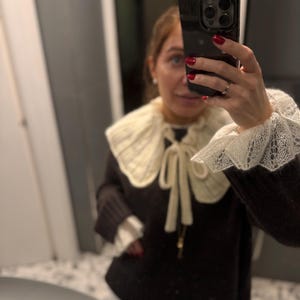 May include: A dark brown sweater with a cream-colored, knitted collar and cuffs. The collar is tied with a cream-colored ribbon. The cuffs have a lace trim. The sweater is worn over a white shirt.