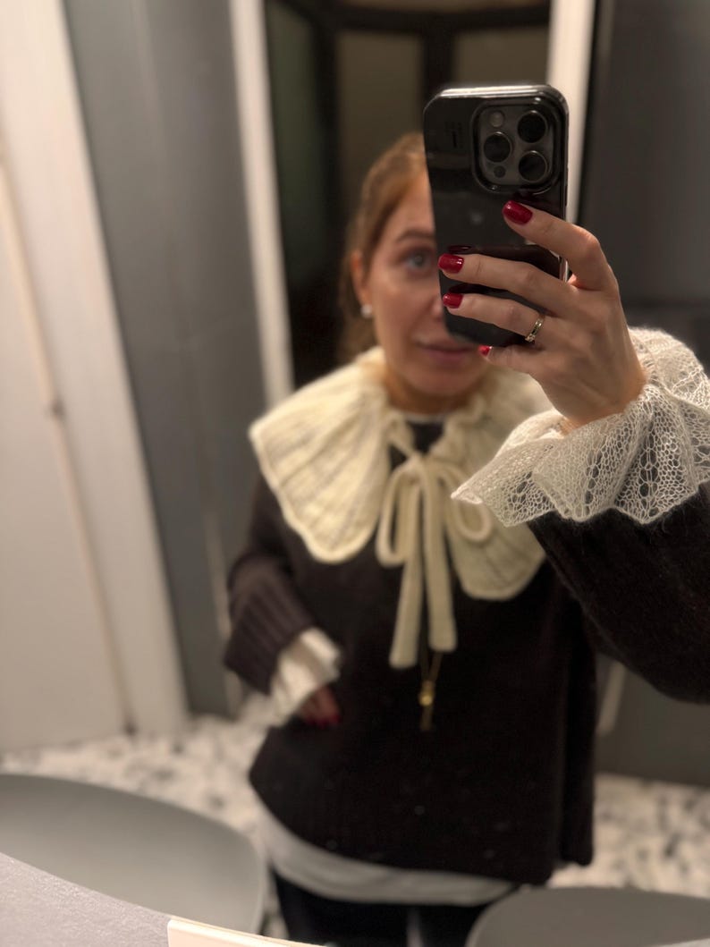 May include: A dark brown sweater with a cream-colored, knitted collar and cuffs. The collar is tied with a cream-colored ribbon. The cuffs have a lace trim. The sweater is worn over a white shirt.