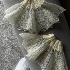 May include: Two off-white, delicate, knitted lace cuffs. The cuffs have a ribbed, cream-colored band at the top and a flared, scalloped edge. The lace has an open, airy design. The cuffs are displayed on a gray surface.