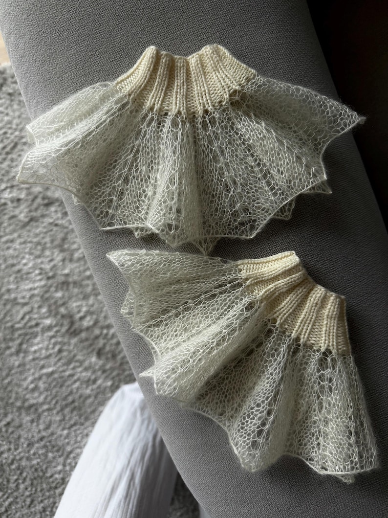 May include: Two off-white, delicate, knitted lace cuffs. The cuffs have a ribbed, cream-colored band at the top and a flared, scalloped edge. The lace has an open, airy design. The cuffs are displayed on a gray surface.