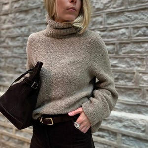 Puede incluir: Una persona lleva un jersey de cuello alto beige, pantalones de pana marrón oscuro y un cinturón de cuero marrón con hebilla dorada. También lleva un anillo plateado y un bolso de ante marrón oscuro.