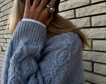 May include: A light blue cable-knit sweater with a textured pattern, worn with a dark gray knit beanie and a clear acrylic ring featuring embedded silver-toned spheres. The sweater has a ribbed collar and cuffs, and the ring is worn on the finger.