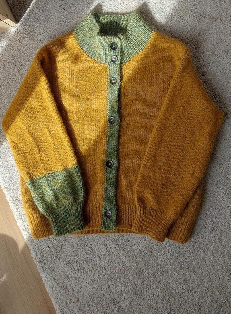 May include: Mustard yellow cardigan with a high collar and button-up front. The collar, button strip, and sleeve cuffs are a contrasting green. The sleeves are long and the cardigan is made of a textured knit.