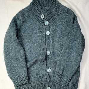 May include: A teal-colored, hand-knitted cardigan sweater with a high collar and five light blue buttons. The sweater has a ribbed collar, cuffs, and hem. The knitwear is a medium weight, suitable for cooler weather.