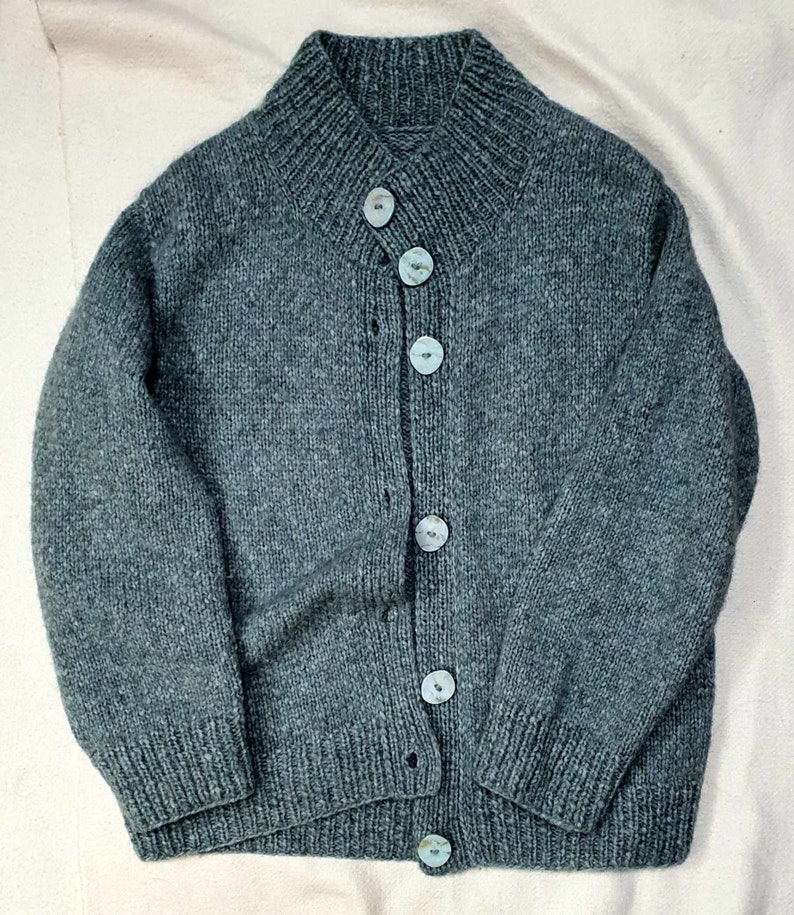May include: A teal-colored, hand-knitted cardigan sweater with a high collar and five light blue buttons. The sweater has a ribbed collar, cuffs, and hem. The knitwear is a medium weight, suitable for cooler weather.