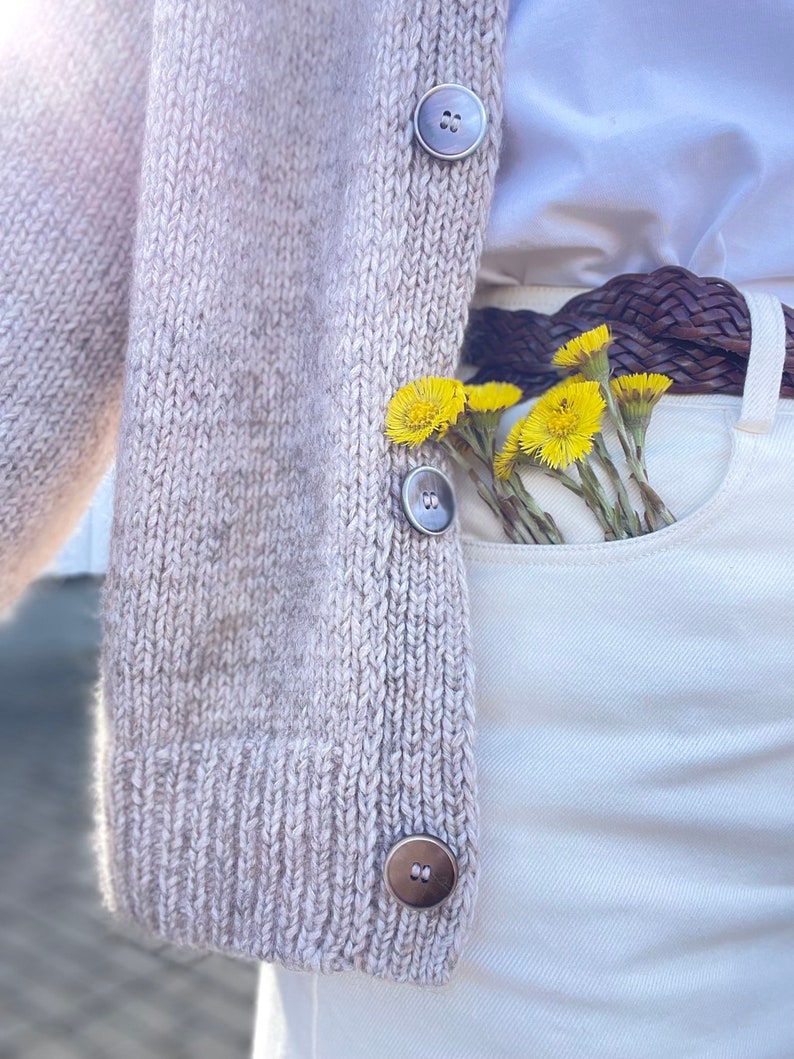May include: A light beige knit cardigan with large, round buttons. The cardigan is paired with white pants, a brown belt, and a white shirt. Yellow flowers are tucked into the pocket, adding a pop of color.