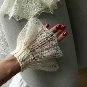 Pode incluir: Luvas de malha rendada de cor marfim com punhos esvoaçantes e com babados. As luvas têm um punho canelado e um delicado padrão rendado na mão e na babado. A imagem mostra um close-up das luvas.