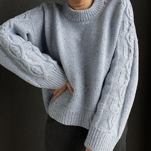 May include: A light blue knitted sweater with a cable knit design on the sleeves. The sweater has a round neckline and a relaxed fit.