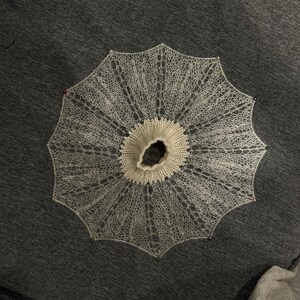 May include: A delicate, off-white, circular knitted lace doily or collar, pinned to a dark gray surface. The intricate openwork design radiates outwards from a central, slightly raised, cream-colored ring. Small pins secure the edges.