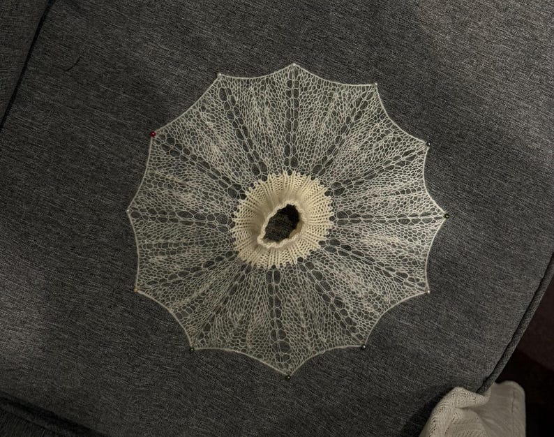 May include: A delicate, off-white, circular knitted lace doily or collar, pinned to a dark gray surface. The intricate openwork design radiates outwards from a central, slightly raised, cream-colored ring. Small pins secure the edges.