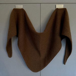 May include: A brown knitted shawl is displayed against a gray background. The shawl is a triangular shape with long, pointed ends. The fabric has a textured appearance, and the color is a warm, earthy brown.