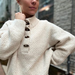 May include: Cream-coloured knitted sweater with a high collar and three wooden toggle buttons. The sweater features a textured pattern and long sleeves. A classic, cosy design, suitable for cooler weather.
