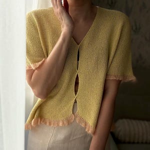 May include: A light yellow, short-sleeved cardigan with a V-neck and a scalloped, peach-colored trim. The cardigan is buttoned at the center and paired with a beige skirt. The outfit is worn by a person standing near a window.