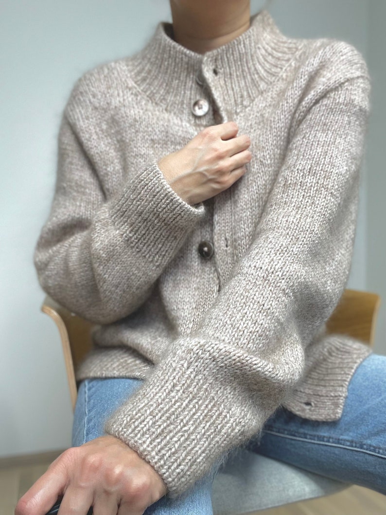May include: A beige, button-down cardigan sweater with a high collar. The sweater is made of a textured knit fabric and features ribbed cuffs and a button closure. The sleeves are long and the sweater appears to be warm and cozy.