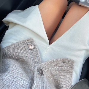 May include: A light gray, chunky knit cardigan with large, round, light-colored buttons. The cardigan is worn over a pair of off-white pants. The image is taken from a first-person perspective, with the steering wheel visible.