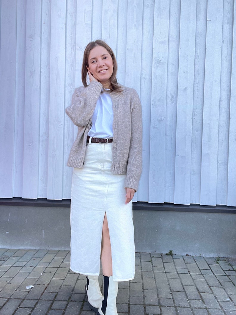 May include: A woman wearing a beige cardigan over a white t-shirt, paired with a cream-colored denim skirt with a front slit. She is also wearing a brown belt and white boots with black accents. The outfit is casual and stylish.