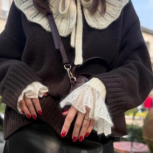 May include: A dark brown sweater with a cream-colored, lace-trimmed collar and cuffs. The sweater has a cream-colored bow at the neckline. A brown purse with a dark brown strap is visible. The person's nails are painted red.
