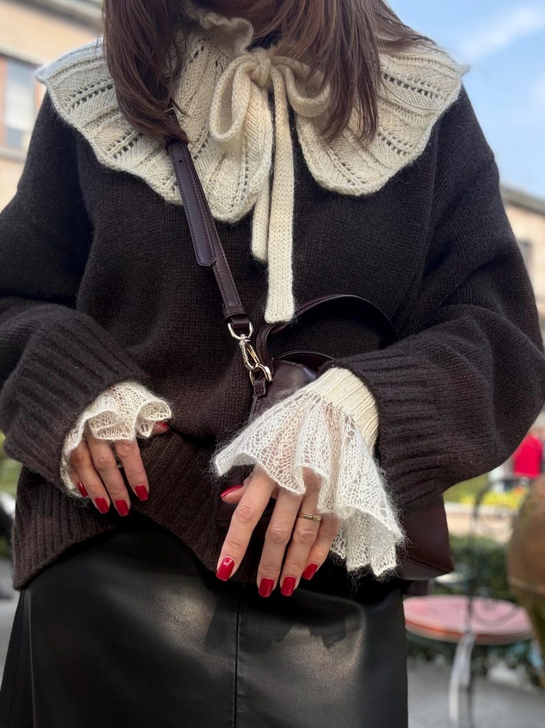 May include: A dark brown sweater with a cream-colored, lace-trimmed collar and cuffs. The sweater has a cream-colored bow at the neckline. A brown purse with a dark brown strap is visible. The person's nails are painted red.