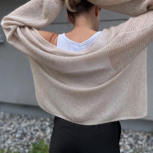 Könnte beinhalten: Ein beigefarbener Strickcardigan mit lockerer Passform und langen Ärmeln. Der Cardigan ist aus einem leichten, luftigen Stoff gefertigt und hat eine leicht verkürzte Länge.