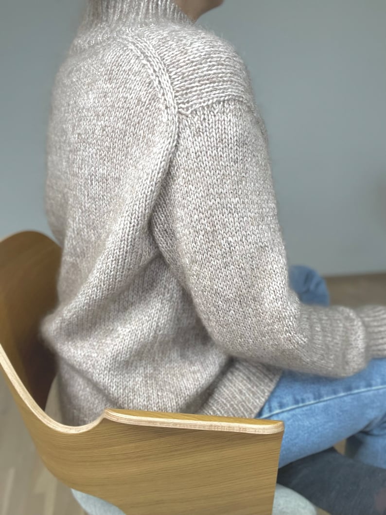 May include: A light beige knit sweater with a high neckline. The sweater has a textured appearance and long sleeves. The image shows the sweater on a person sitting in a wooden chair. The sweater is paired with blue jeans.
