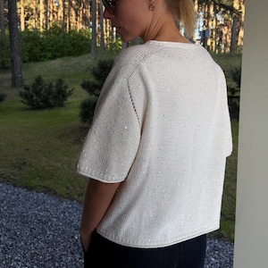 Könnte beinhalten: Cremefarbener Pullover mit kurzen Ärmeln und dezenten Paillettenverzierungen. Das Strickoberteil hat einen runden Ausschnitt und eine leicht verkürzte Passform. Der Pullover wird mit einer dunklen Hose kombiniert, was einen lässigen Look ergibt.