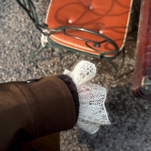May include: A brown jacket with a dark brown cuff and a white, open-weave lace sleeve. The jacket is buttoned and the sleeve is flared. An orange chair is in the background.