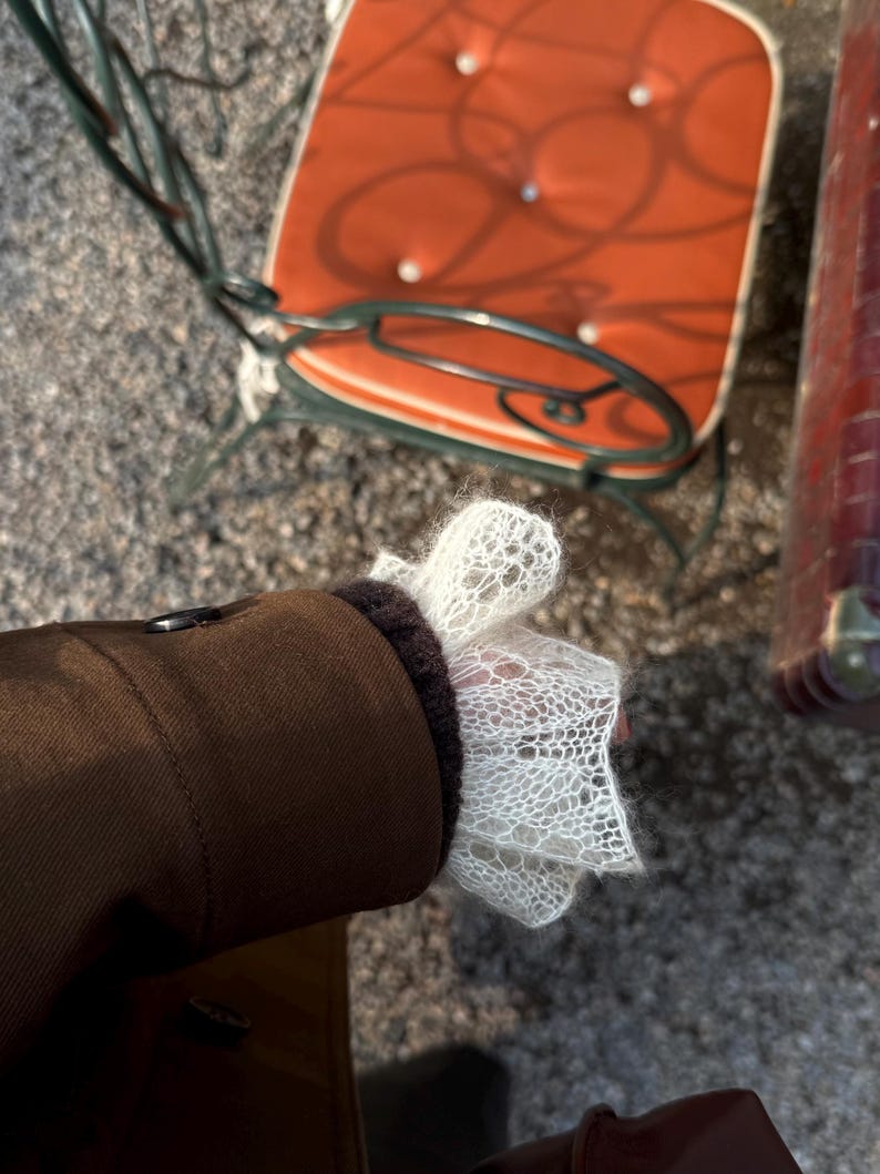 May include: A brown jacket with a dark brown cuff and a white, open-weave lace sleeve. The jacket is buttoned and the sleeve is flared. An orange chair is in the background.