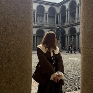 May include: A person wearing a brown coat with a white collar and cuffs, standing in a courtyard. The coat is paired with a long, dark skirt. The background features a building with arches and statues, framed by stone columns.