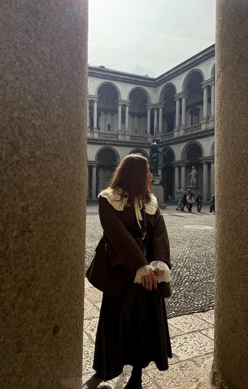 May include: A person wearing a brown coat with a white collar and cuffs, standing in a courtyard. The coat is paired with a long, dark skirt. The background features a building with arches and statues, framed by stone columns.