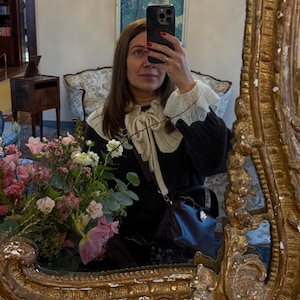 May include: A woman taking a mirror selfie, framed by a gold ornate mirror. She wears a black sweater with a white lace collar and cuffs. A small, dark brown handbag with a silver clasp hangs from her shoulder. A bouquet of pink and white flowers is visible.