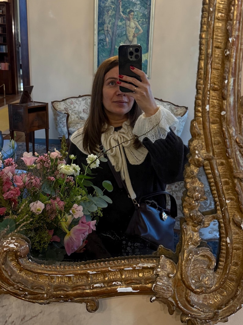 May include: A woman taking a mirror selfie, framed by a gold ornate mirror. She wears a black sweater with a white lace collar and cuffs. A small, dark brown handbag with a silver clasp hangs from her shoulder. A bouquet of pink and white flowers is visible.