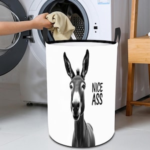 May include: A black and white laundry hamper with a donkey graphic and the text "NICE ASS". The hamper has black handles and is next to a washing machine. A person is putting clothes into the washing machine.