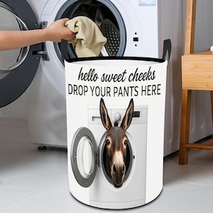 Puede incluir: Una cesta de lavandería blanca con asas negras y el texto "hello sweet cheeks DROP YOUR PANTS HERE". La cesta presenta la cabeza de un burro que emerge de una lavadora.