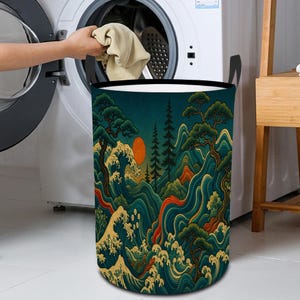 Pode incluir: Um cesto de roupa cilíndrico com um design de paisagem em estilo japonês. A obra de arte apresenta ondas do oceano, árvores, montanhas e um sol poente em tons de azul, verde e laranja. O cesto tem alças pretas e um interior branco.
