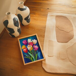 May include: A framed painting of colorful tulips with a blue background rests on a wooden floor. Two white vases with black abstract designs are nearby. A beige rug with abstract shapes is also visible.