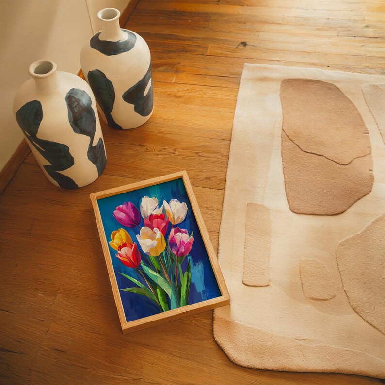 May include: A framed painting of colorful tulips with a blue background rests on a wooden floor. Two white vases with black abstract designs are nearby. A beige rug with abstract shapes is also visible.