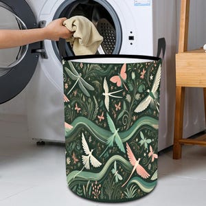 May include: A dark green laundry hamper with a repeating pattern of stylized dragonflies, birds, and butterflies in white, pink, and teal. The hamper has black handles and is next to a washing machine.
