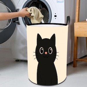 Puede incluir: Una cesta de lavandería de color crema con una ilustración de un gato negro. El gato tiene ojos grandes y redondos, bigotes y una nariz rosa. La cesta tiene asas negras y está junto a una lavadora.