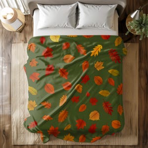 May include: A green blanket featuring a pattern of autumn leaves in red, orange, and yellow. The leaves are scattered across the blanket, creating a fall-themed design. The blanket is on a bed with white pillows.