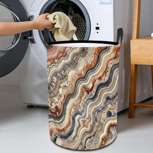 May include: A cylindrical laundry hamper with a decorative pattern of swirling bands in shades of brown, beige, and red. The hamper has black handles and a black rim. A person is putting clothes into a washing machine.