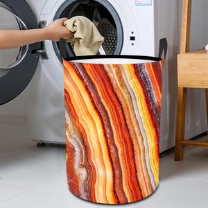 May include: A cylindrical laundry basket featuring a colourful, abstract pattern in shades of orange, yellow, and brown, resembling layered agate stone. It has black handles and a black rim.