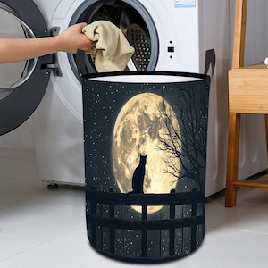May include: A black laundry hamper with a moon and cat silhouette design. The moon is full and bright, with a starry night sky in the background. The cat is sitting on a fence, looking up at the moon.