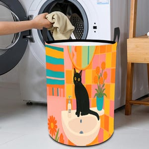 Könnte beinhalten: Ein runder Wäschekorb mit einer farbenfrohen Illustration einer schwarzen Katze, die auf einem Waschbecken sitzt. Der Hintergrund zeigt eine geflieste Wand mit orangefarbenen, rosa und gelben Quadraten. Der Korb hat schwarze Griffe.